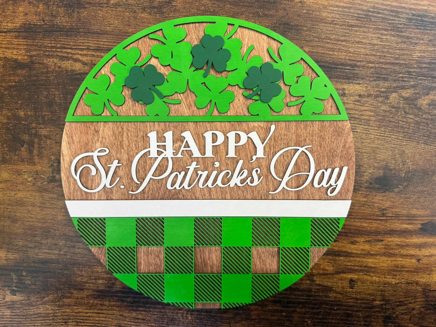 A round decorative sign with a plaid pattern and clover leaves at the top, featuring the text 'HAPPY St. Patrick's Day' in the center, displayed on a wooden surface.