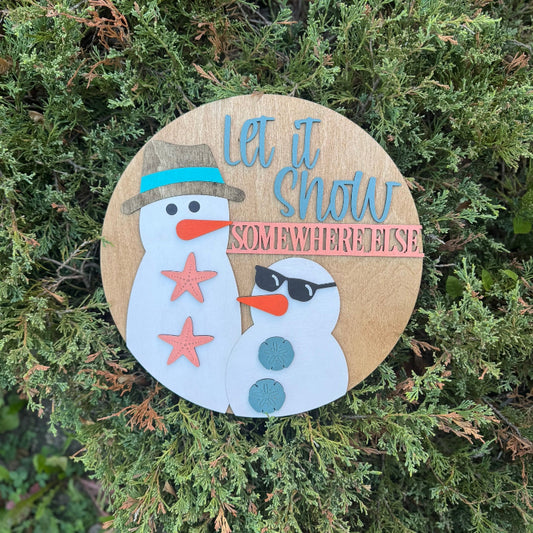 Round wooden sign with a snowman design and 'Let it snow somewhere else' text on a green bush background.