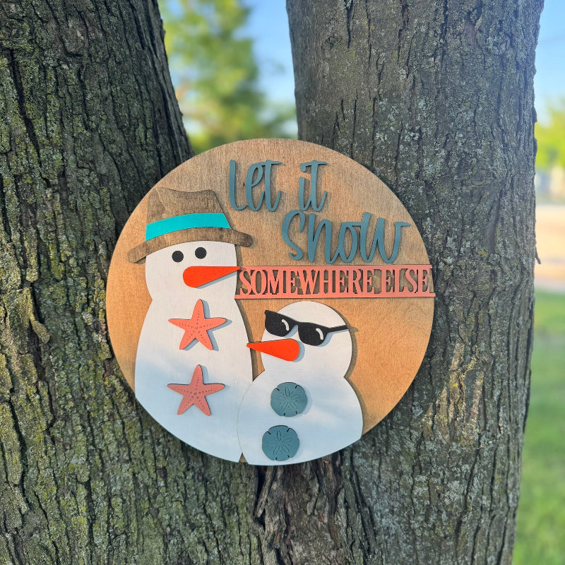Decorative sign with a snowman design and text 'let it snow somewhere else' attached to a tree.