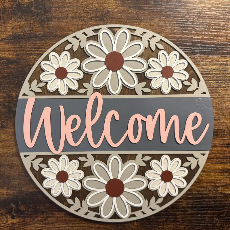 A round decorative sign with a floral pattern and the word 'Welcome' in the center, displayed on a wooden surface.