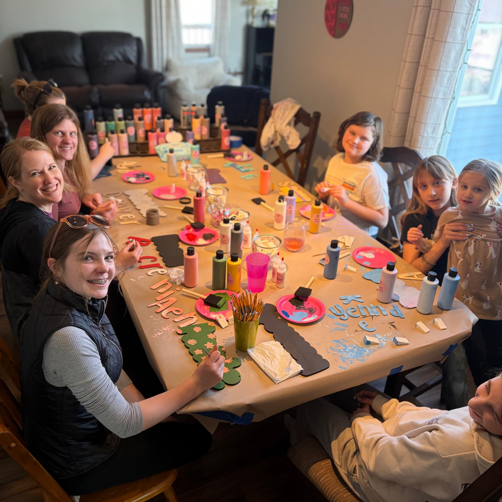Group of people, including children and adults, engaged in a name sign painting activity at a table with various supplies.