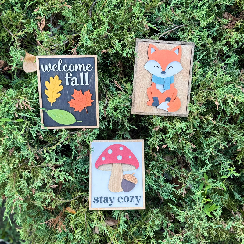 Three wooden signs with autumn-themed designs on a green leafy background.