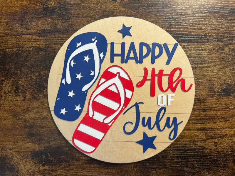 A round wooden sign with 'Happy 4th of July' text, featuring stars and stripes, and a pair of red and blue flip-flops.