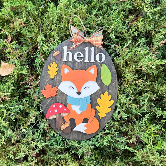 Decorative sign with a fox illustration and 'hello' text, surrounded by autumn leaves and a bow, on a green leafy background.