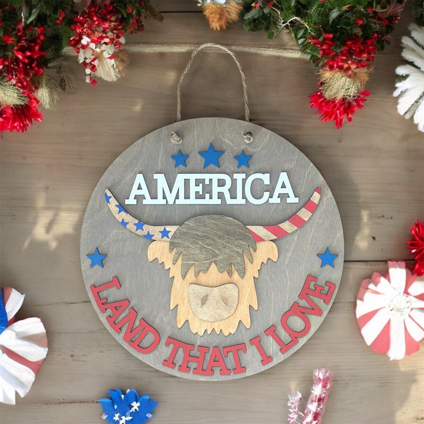 Decorative sign with 'America Land That I Love' text and a Highland Cow head design on a wooden background.