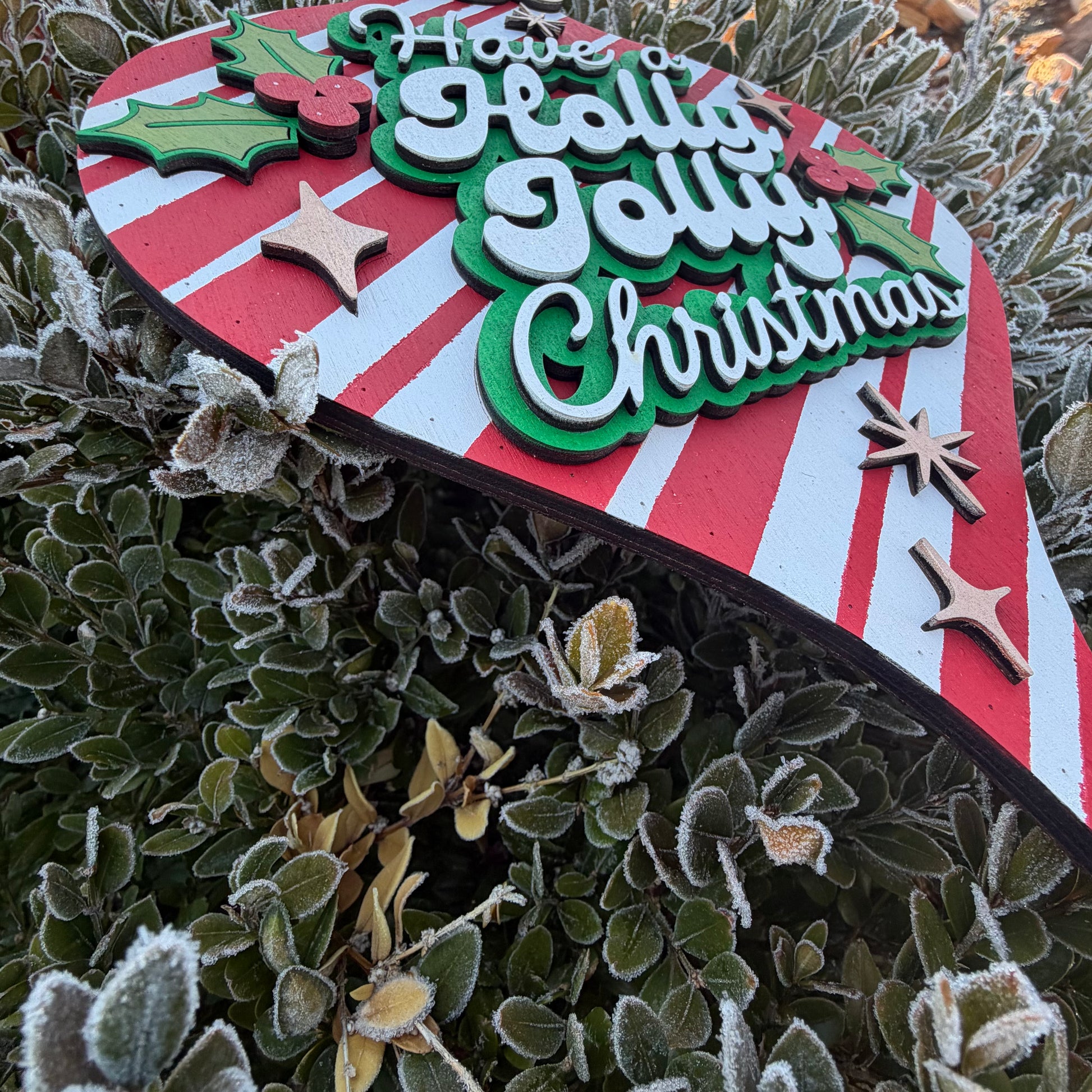 Decorative Christmas sign with 'Have a Holly Jolly Christmas' text on a red and white striped background, surrounded by frosted greenery.