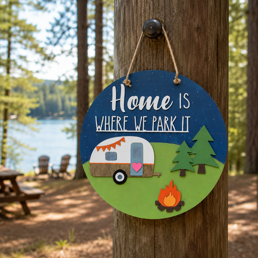 Round wooden hanging sign with the phrase “Home is where we park it” in white lettering on a dark blue background, decorated with a retro camper, green trees, and a campfire on a grassy green section. The sign is shown hanging from a wooden post in a forest campsite setting with a lake in the background.