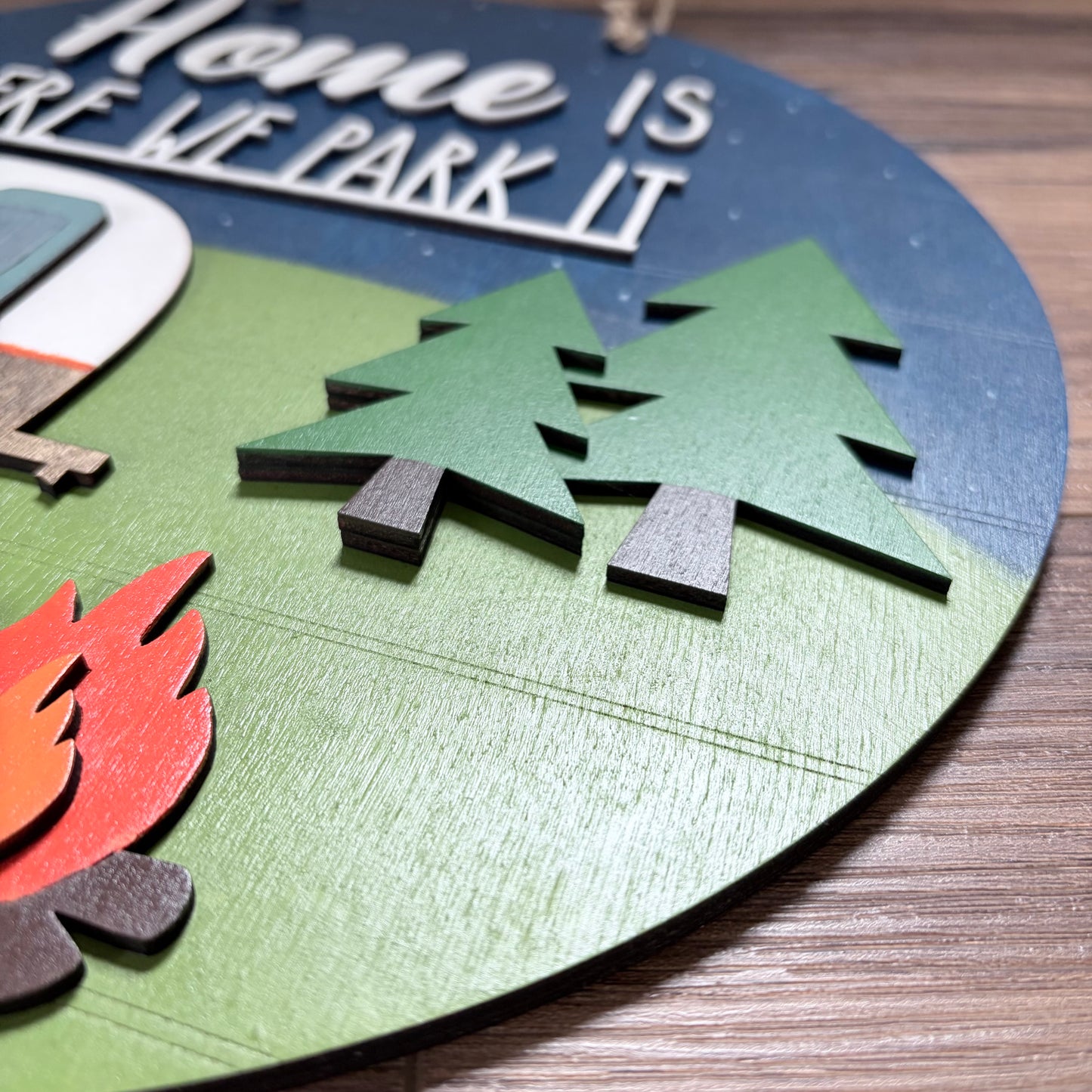A close-up of a round wooden wall sign featuring a camping theme with layered cutouts of pine trees, a campfire, a tent, and a blue sky background. The sign is placed on a wooden tabletop.