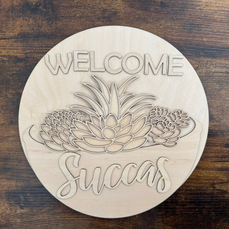 A round wooden welcome sign with a succulent design and the text 'WELCOME Succas' in pink and green colors.