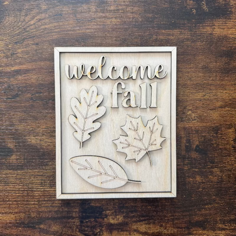 Decorative sign with 'welcome fall' text and leaf designs on a wooden background