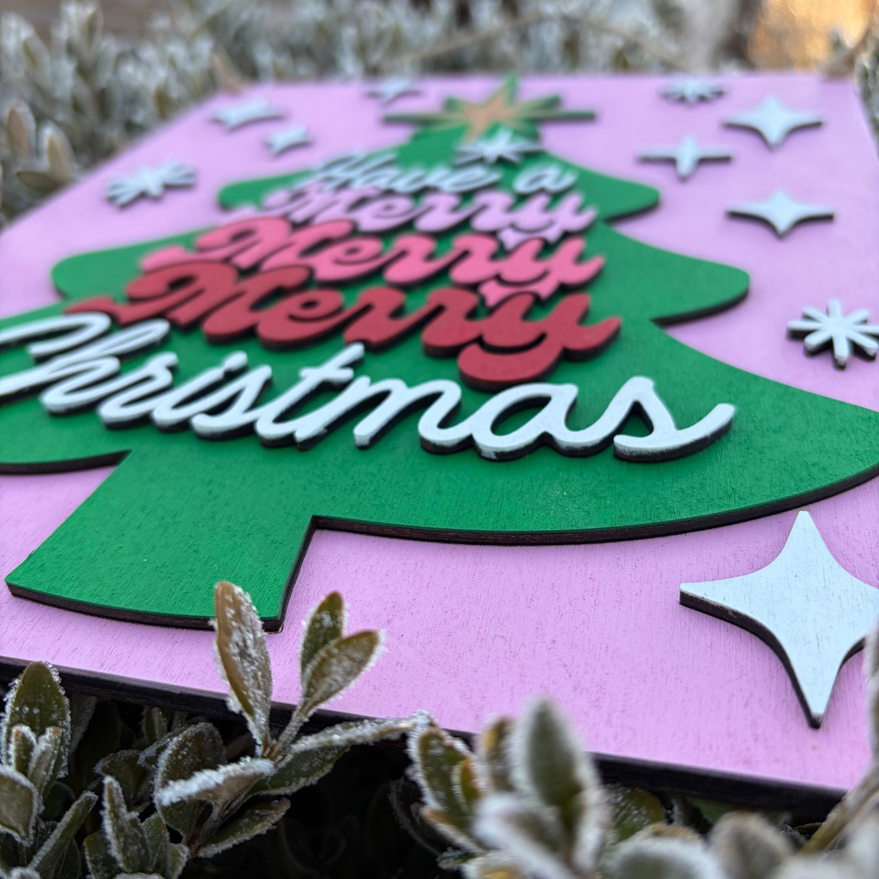 Christmas-themed card with a green tree and 'Merry Christmas' text on a pink background, surrounded by plants.