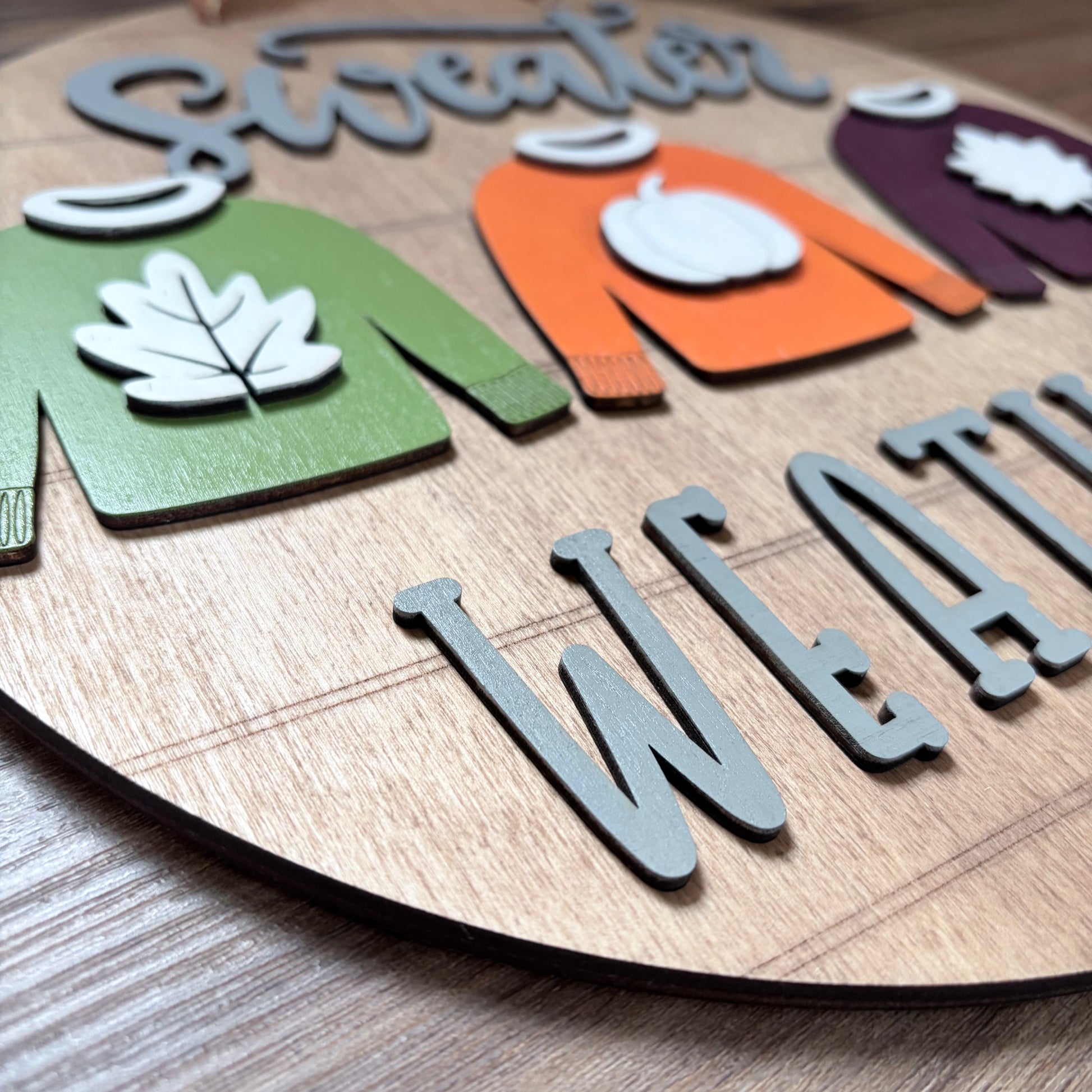 A close-up of a wooden decorative sign featuring stylized autumn sweaters in green, orange, and purple with white leaf and pumpkin details, alongside gray raised letters spelling 'WEATHER'.