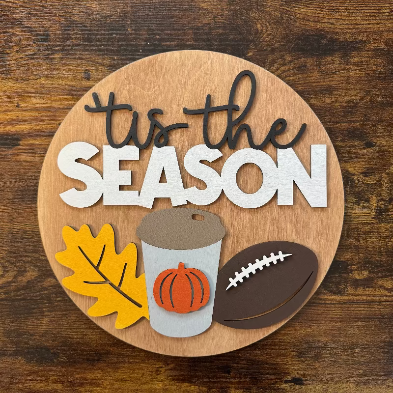 Decorative sign with 'tis the SEASON' text and autumn-themed icons on a wooden background