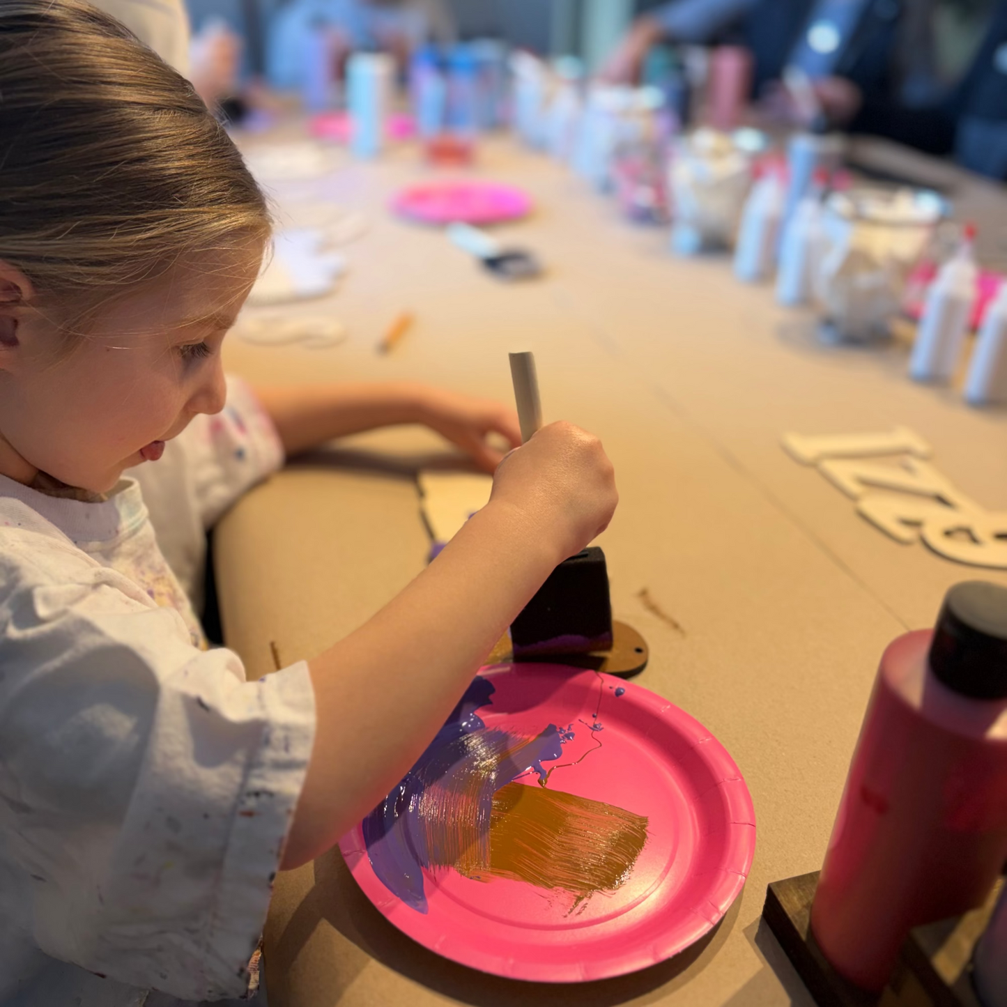 Child painting with a brush on a pink paper plate at a craft activity.