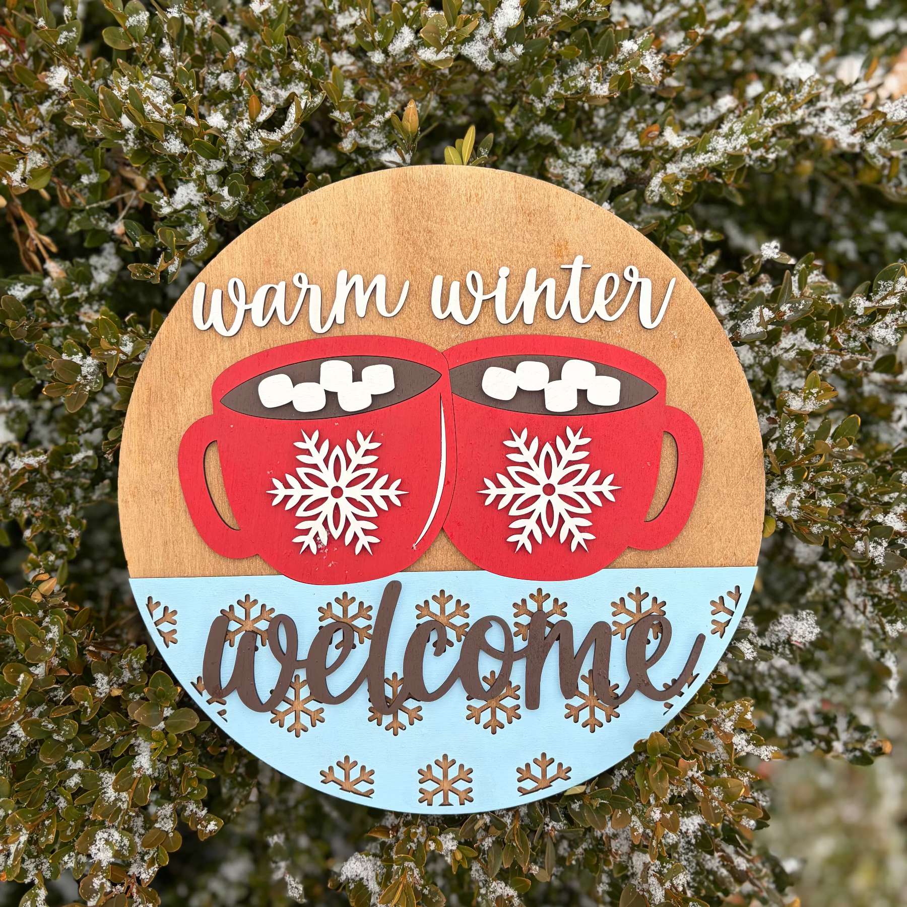 Decorative sign with red mugs, snowflakes, and 'warm winter welcome' text on a wooden background.