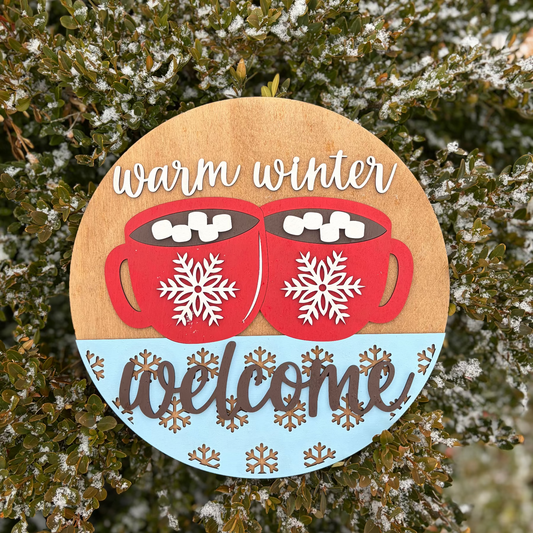 Decorative sign with red mugs, snowflakes, and 'warm winter welcome' text on a wooden background.