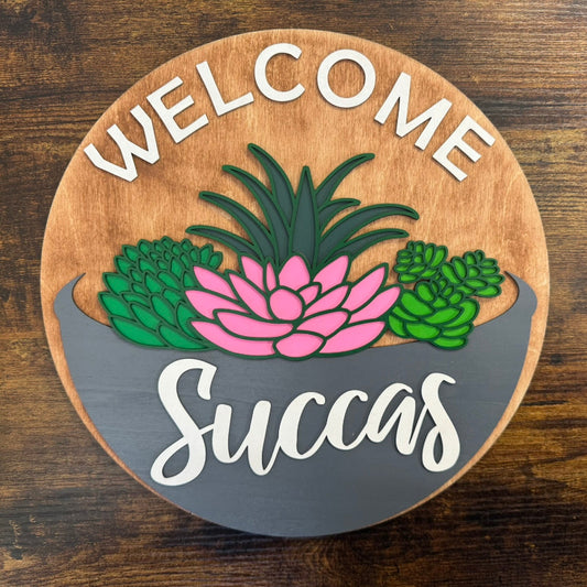 A round wooden welcome sign with a succulent design and the text 'WELCOME Succas' in pink and green colors.
