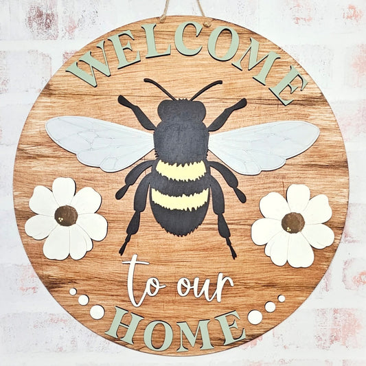 A round wooden welcome sign featuring a bumble bee design with the text 'WELCOME to our HOME' surrounded by floral accents.